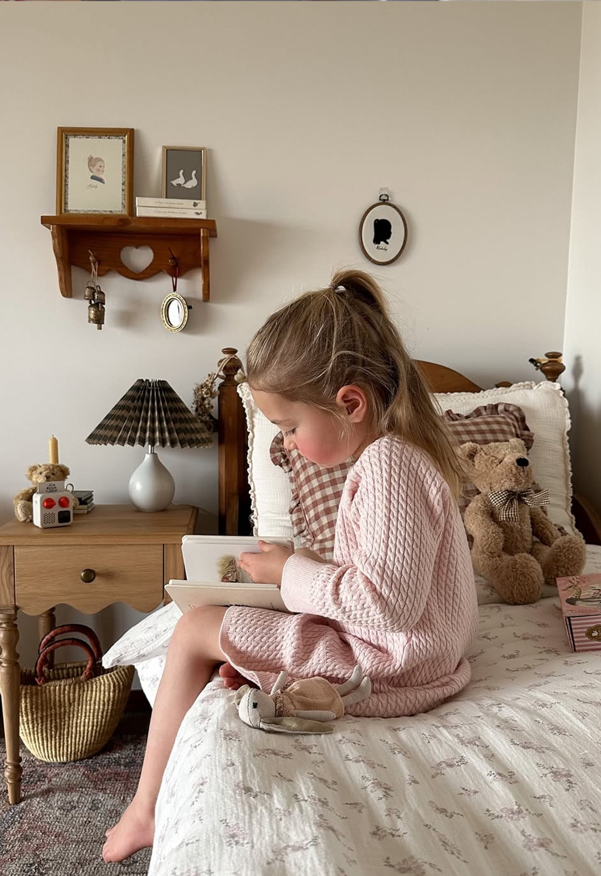 Child in a pink dress sitting on a bed with teddy bears and decorative items in the room.