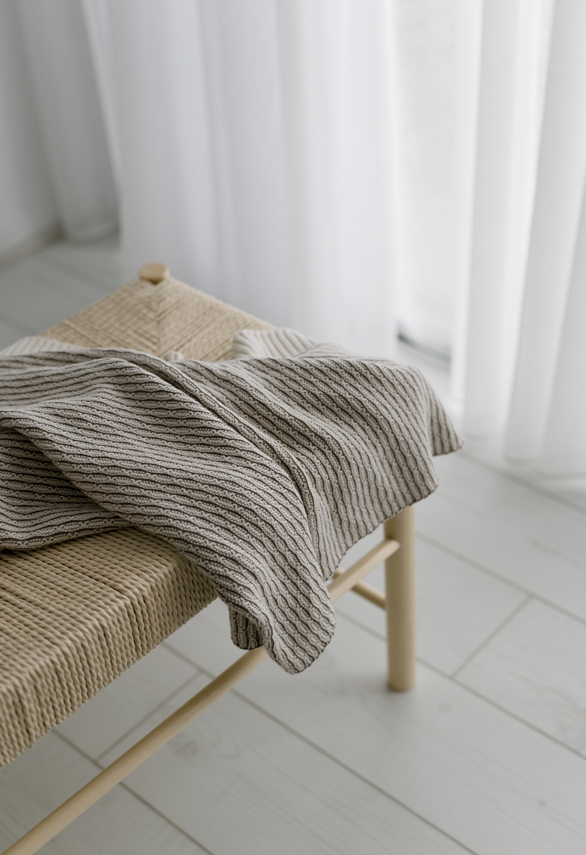 A fay knit structured blanket in shades of beige and brown, draped over a wooden chair with a window in the background.