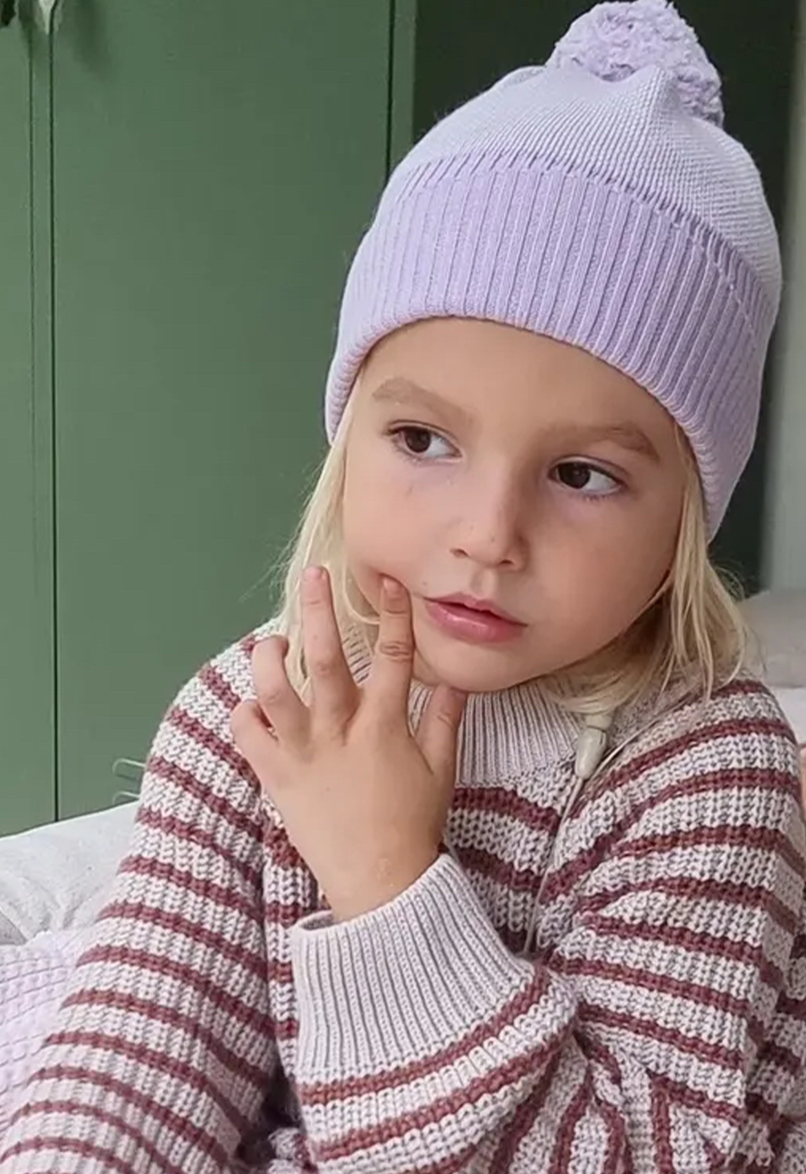 Child wearing a lilac knit hat and striped sweater, sitting against a green background