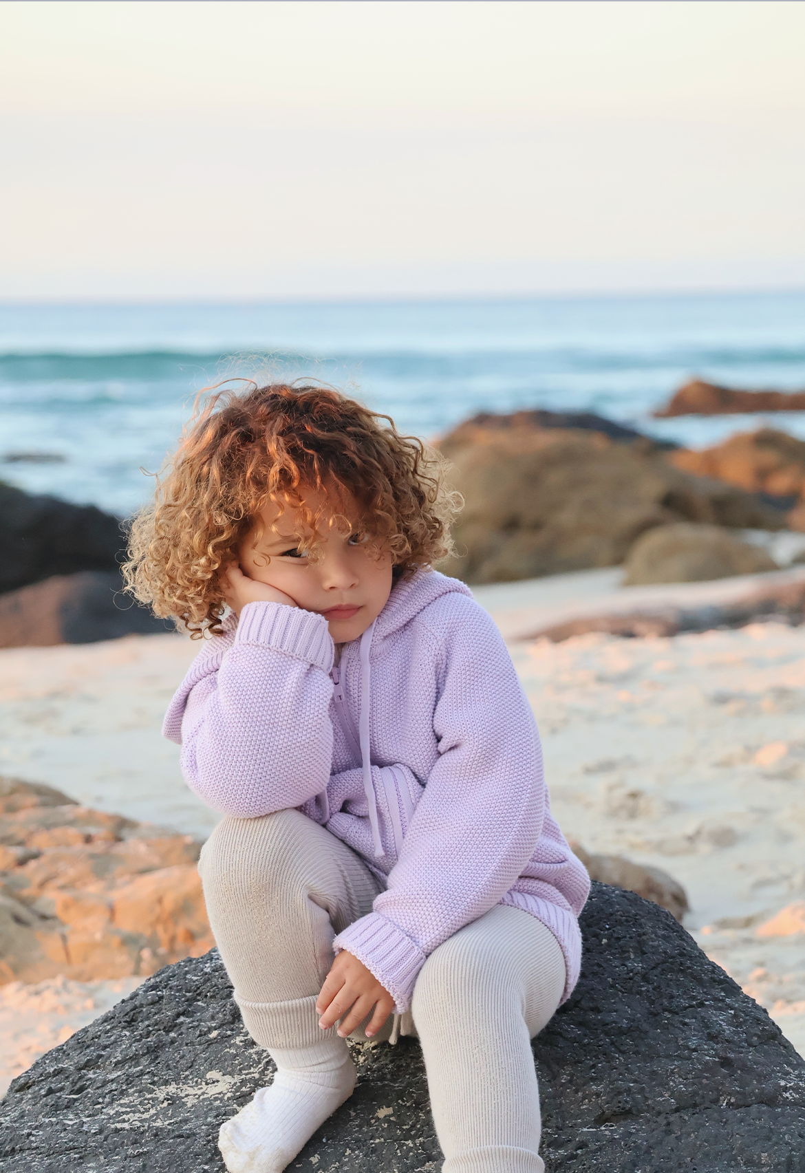 Child wearing a lilac purple hoodie sitting on a rock by the ocean.