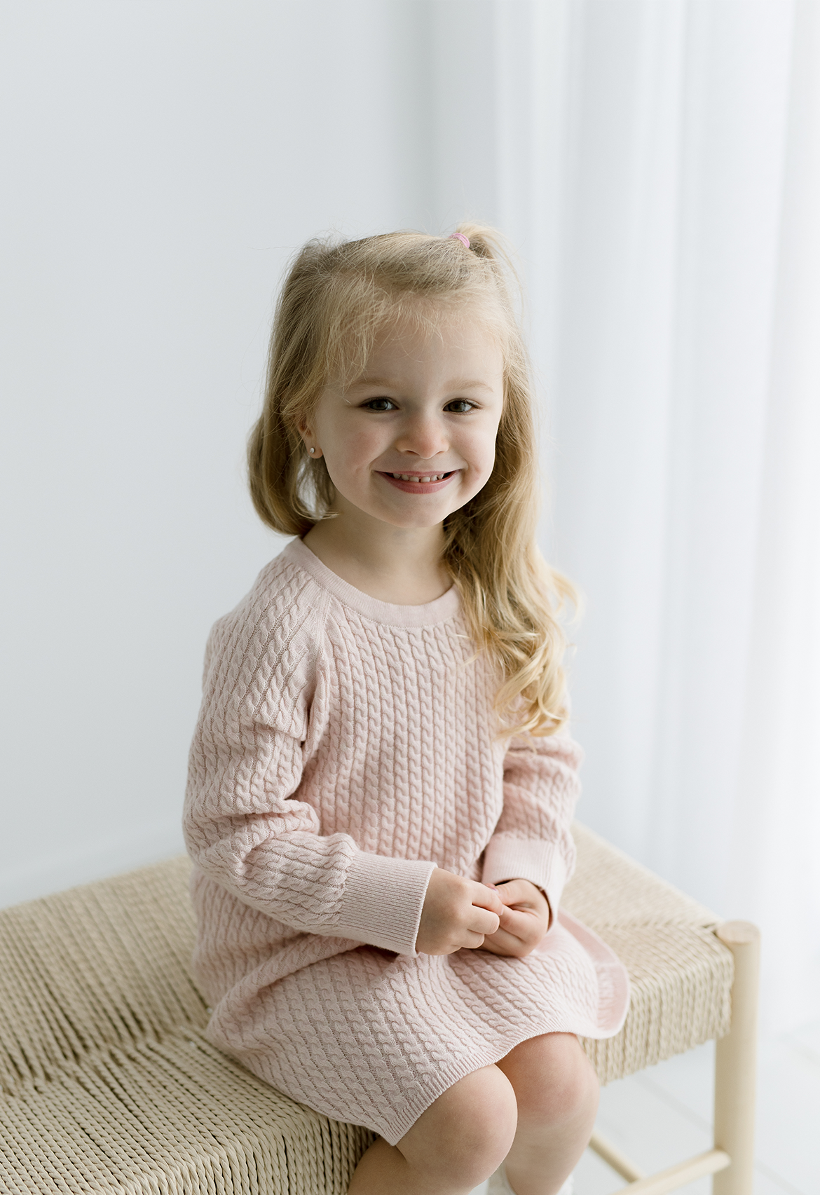A young girl sitting and smiling, wearing a pink cable knit dress.