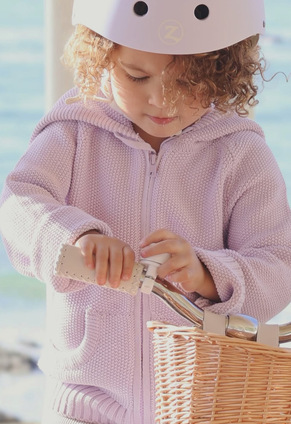 Child in a lilac jacket and helmet sitting in a bicycle basket by a body of water.