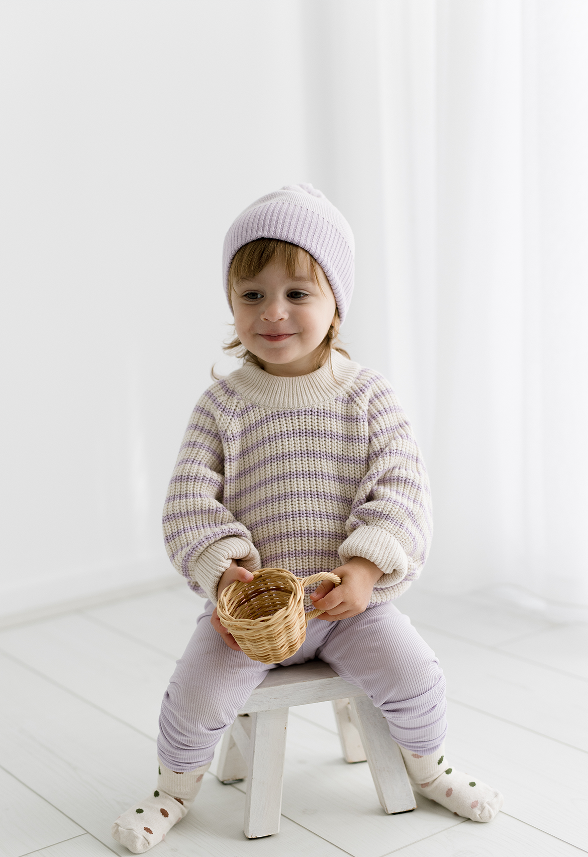 A child sitting on a stool wearing a beige and mauve striped knit jumper with a round neckline.