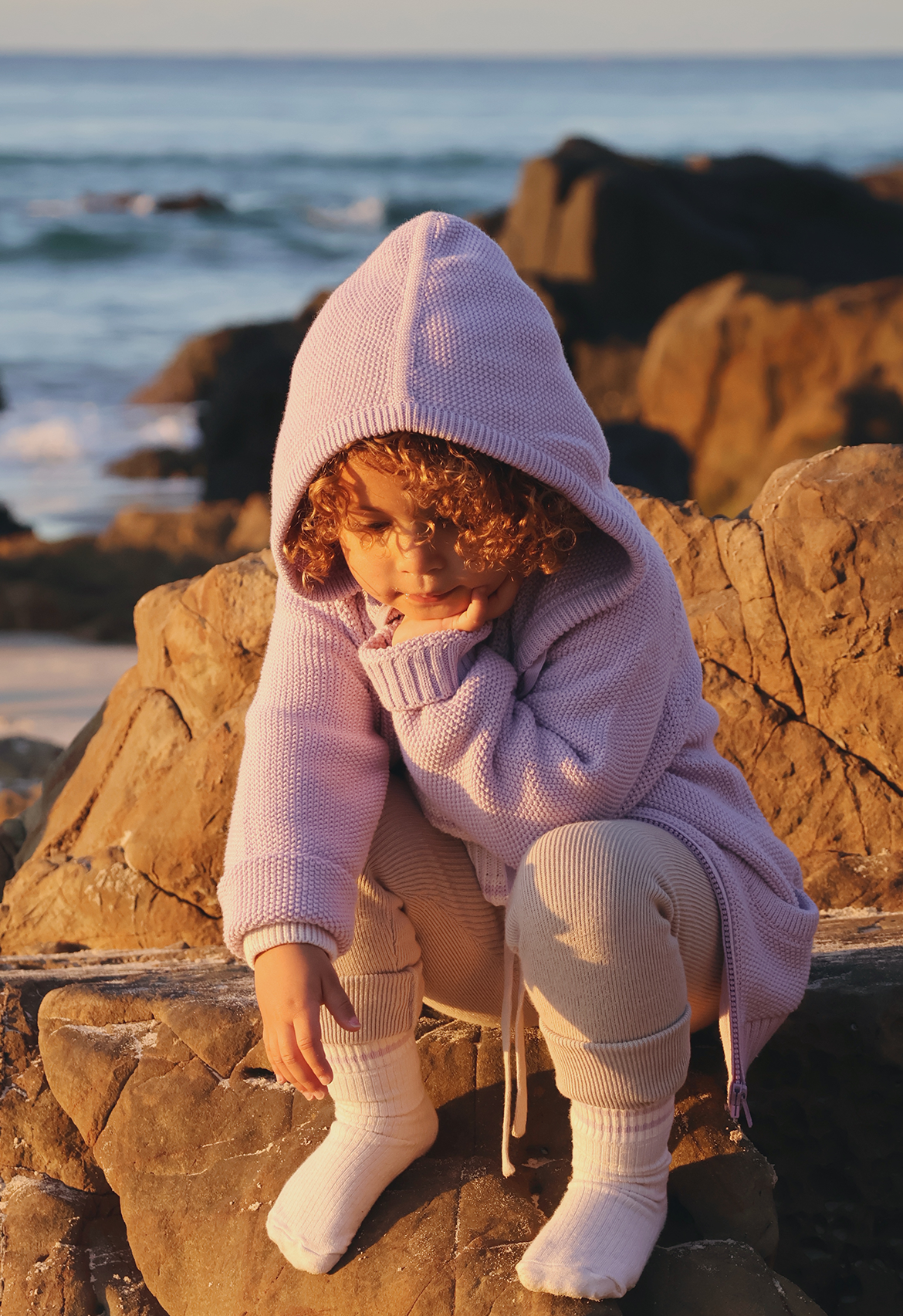 Child in a lilac hooded jacket sitting on rocks by the ocean
