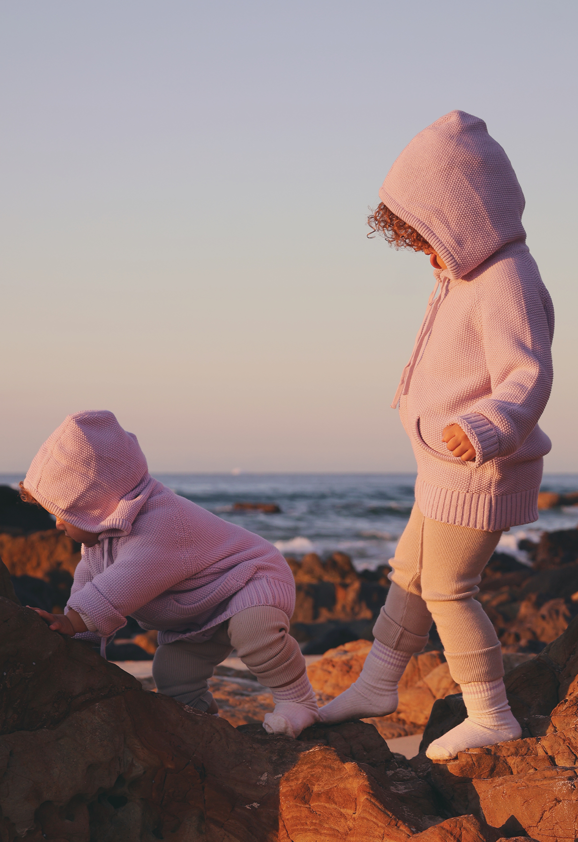 Two children in lilac hoodies standing on rocks by the ocean at sunset.