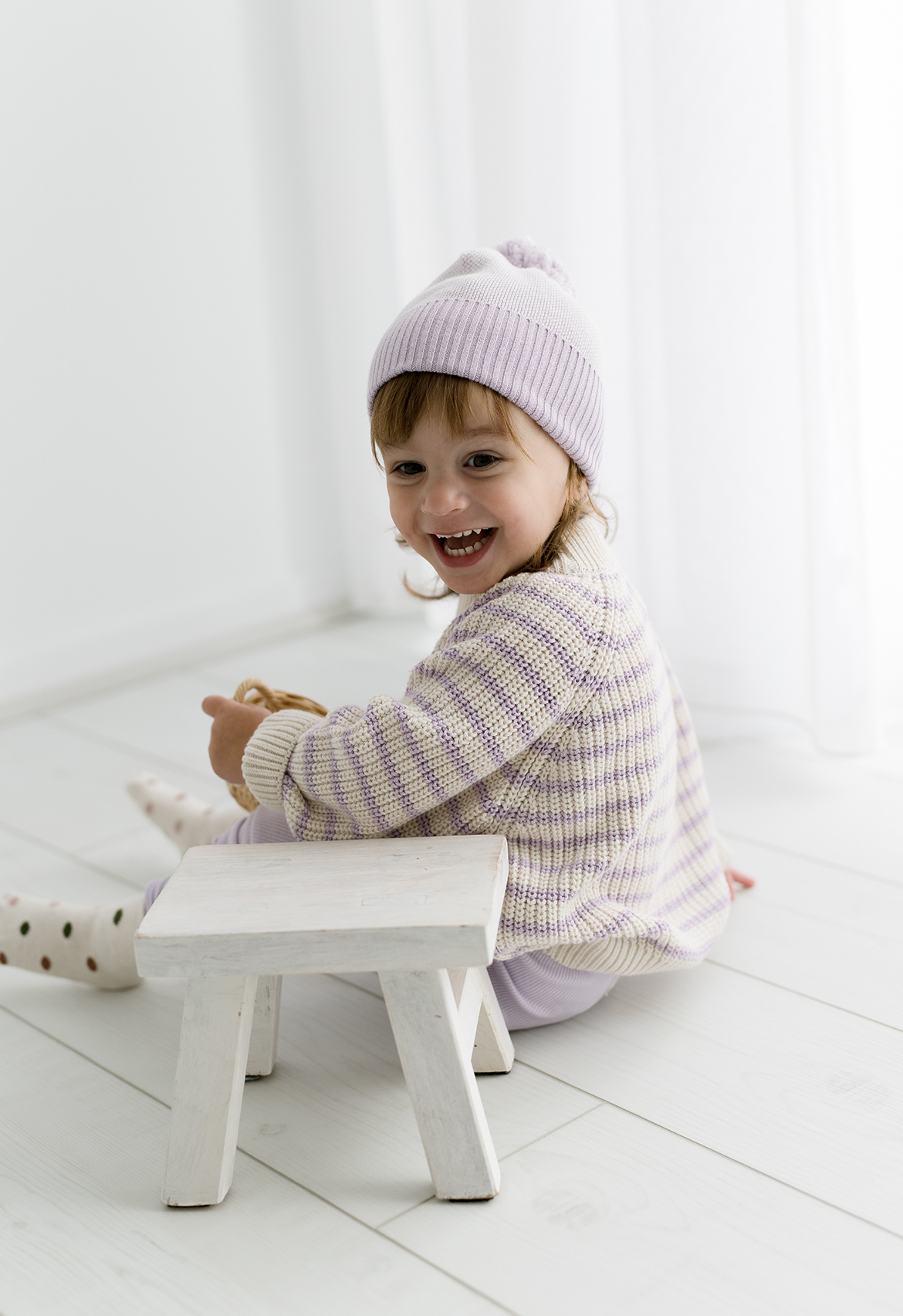 Child wearing a lilac knit beanie sitting on a white stool in a minimalistic room.