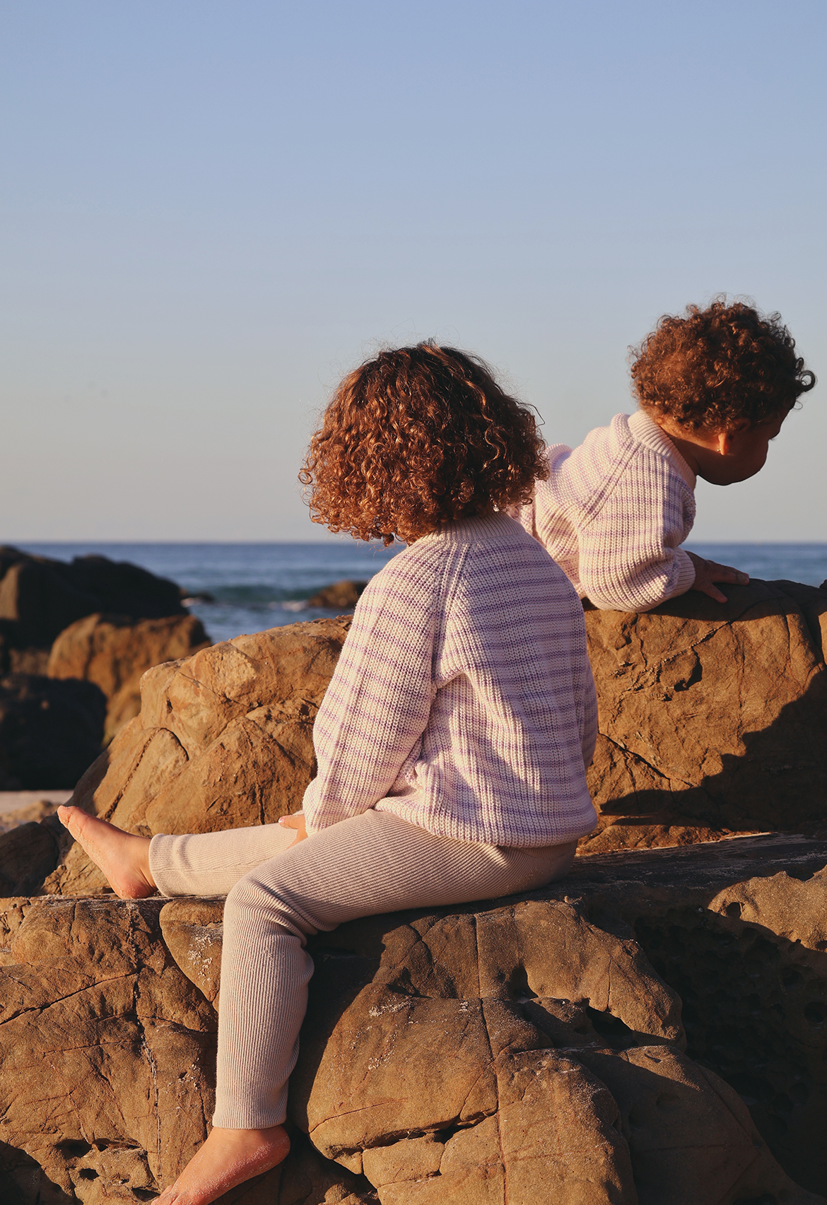Two people sitting on rocks by the ocean at sunset in knitwear.