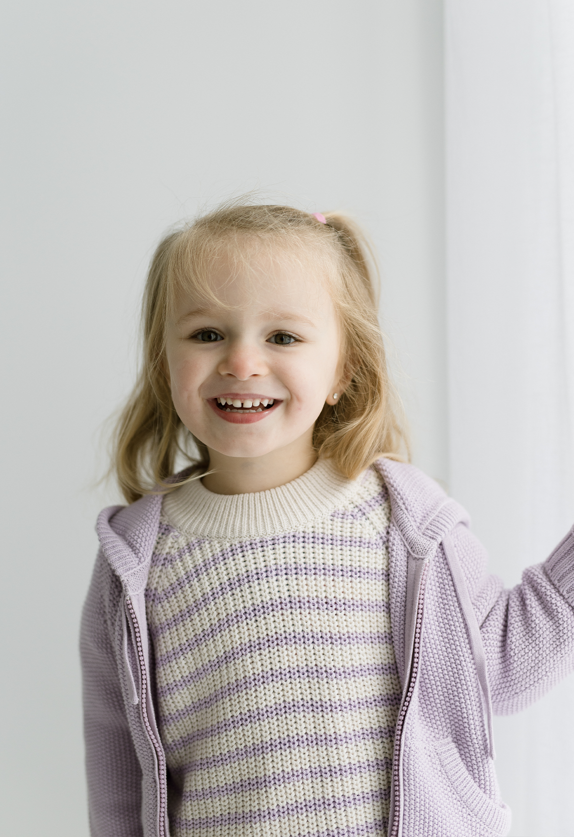 A child smiling and wearing a hooded jacket in lilac color with a zip front, hood with drawstring, and side pockets.