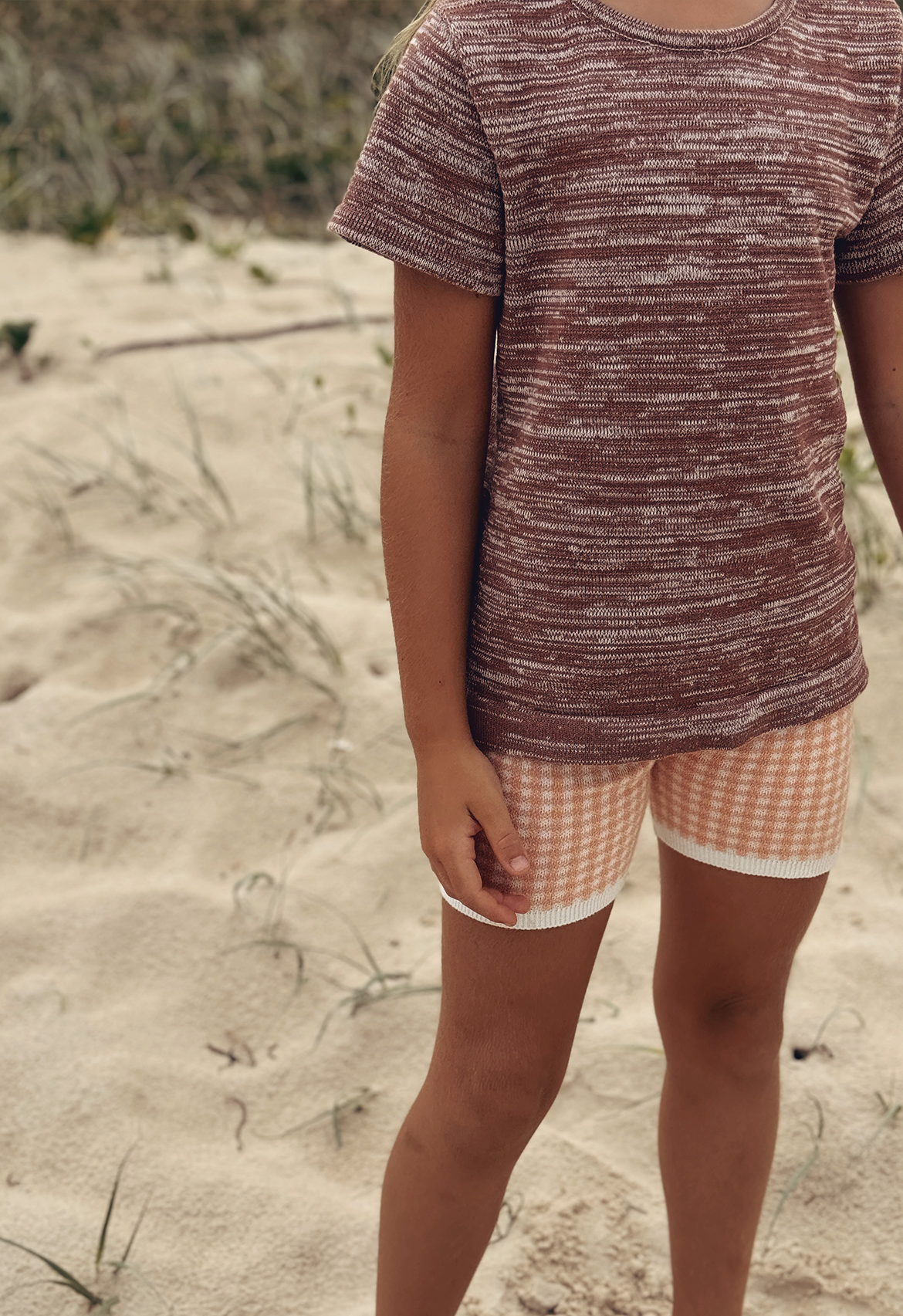 A child standing on a sandy beach wearing a brown, rust textured tee with short sleeves and a round neckline, paired with checkered shorts.