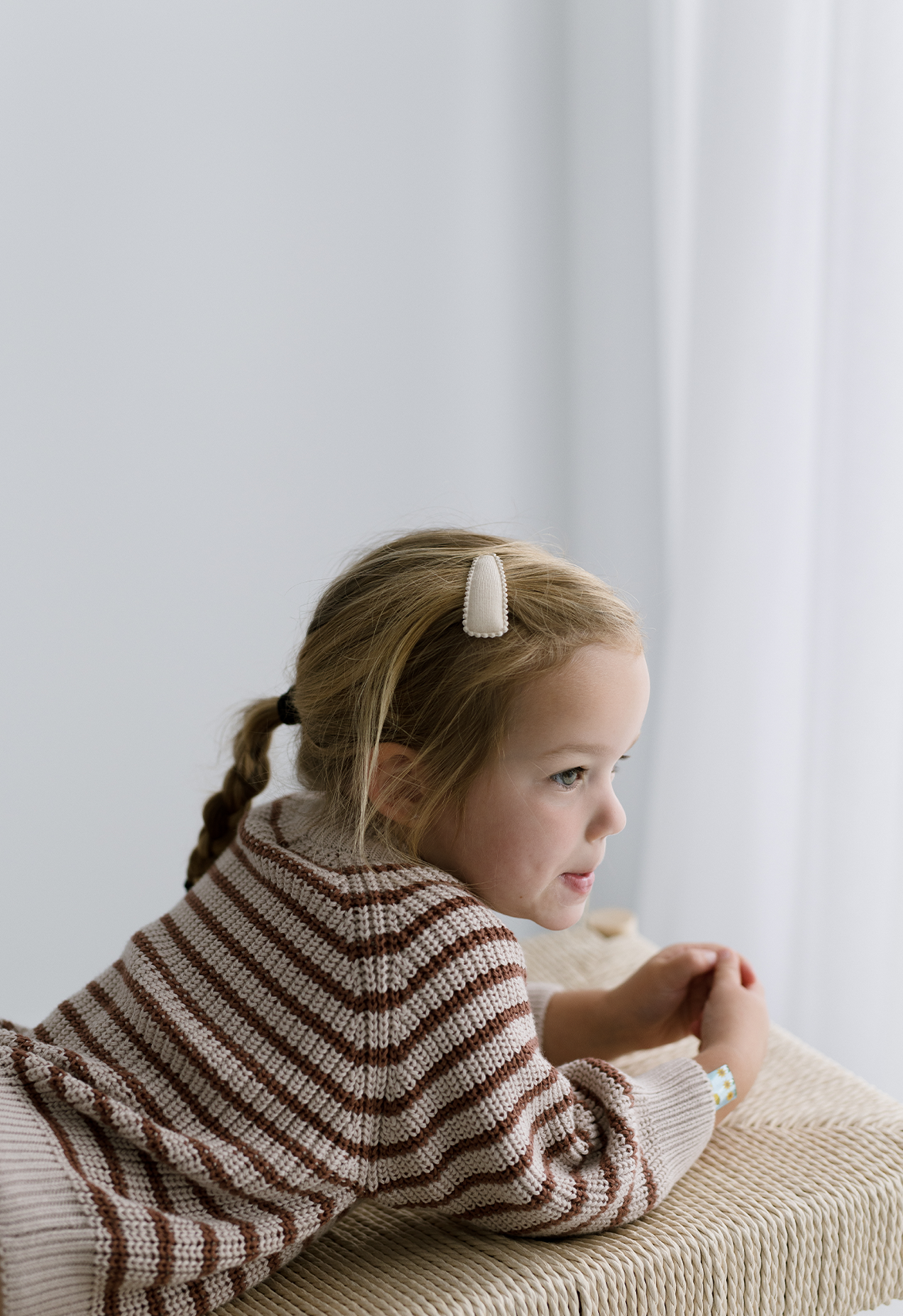 A child wearing a striped jumper in taupe and rust colors, sitting by a window.
