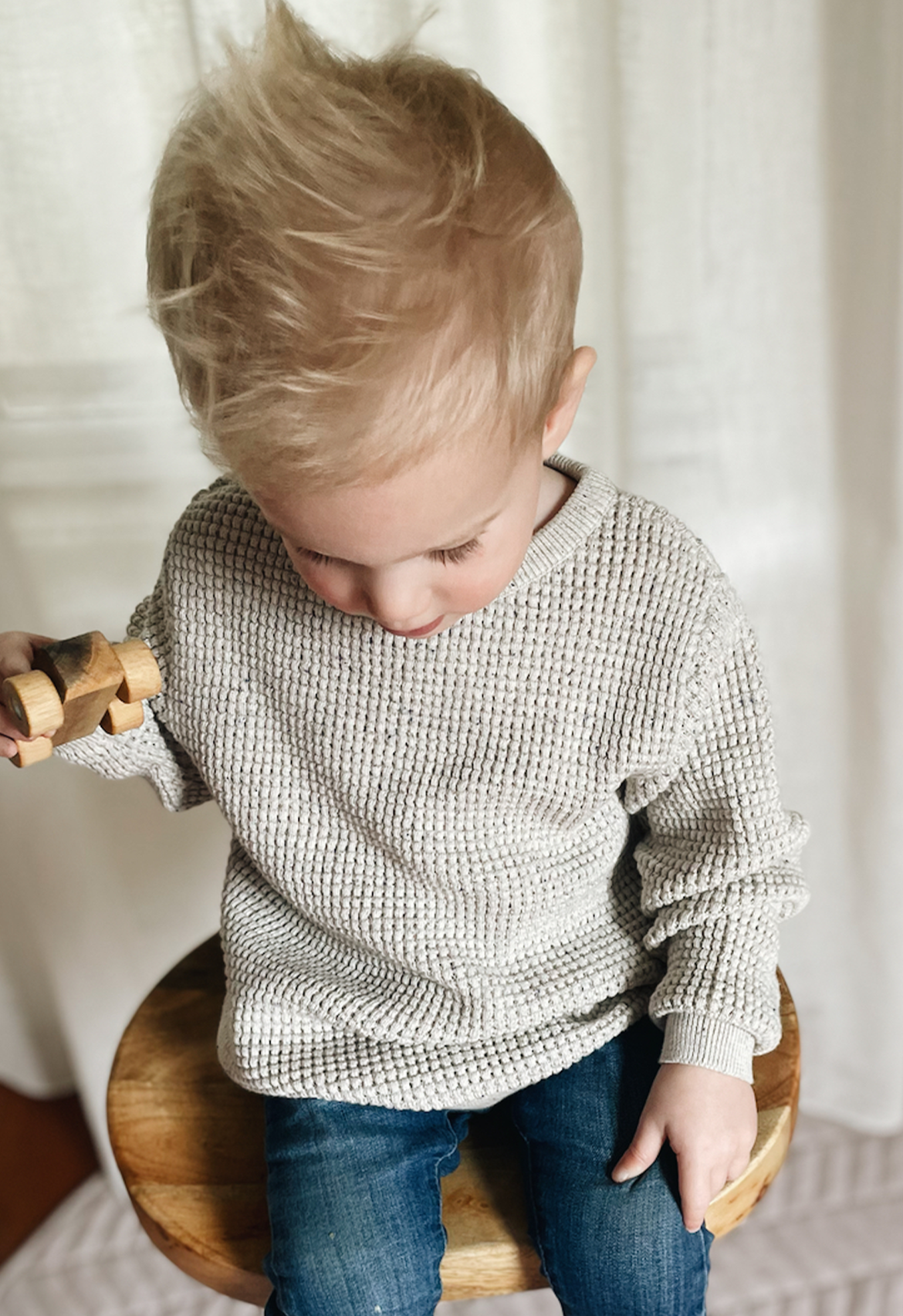toddler sitting on chair wearing waffle speckle jumper, playing with cars matching his jumper with denim jeans
