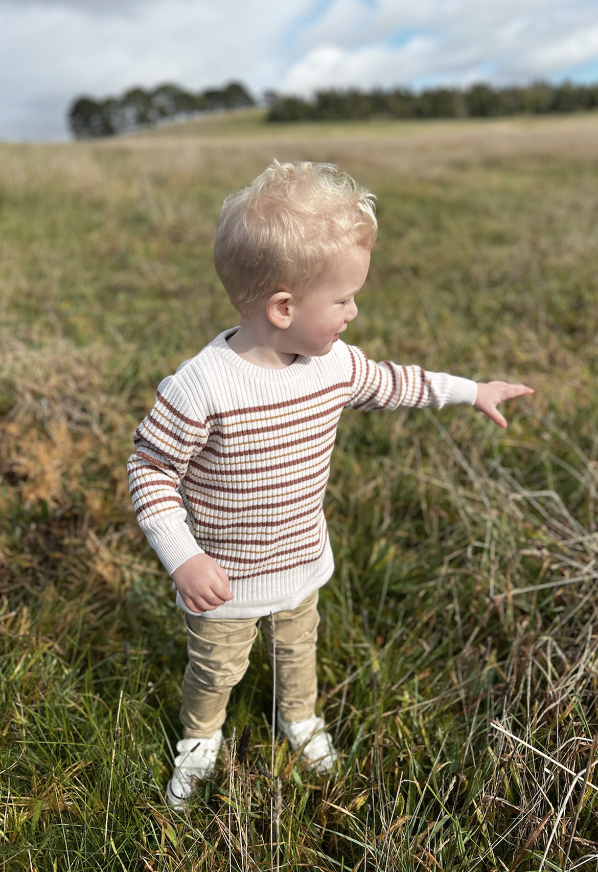 A toddler standing in a field of dry grass, wearing a long sleeve striped jumper in oatmeal with russet and mustard stripes, paired with chinos.