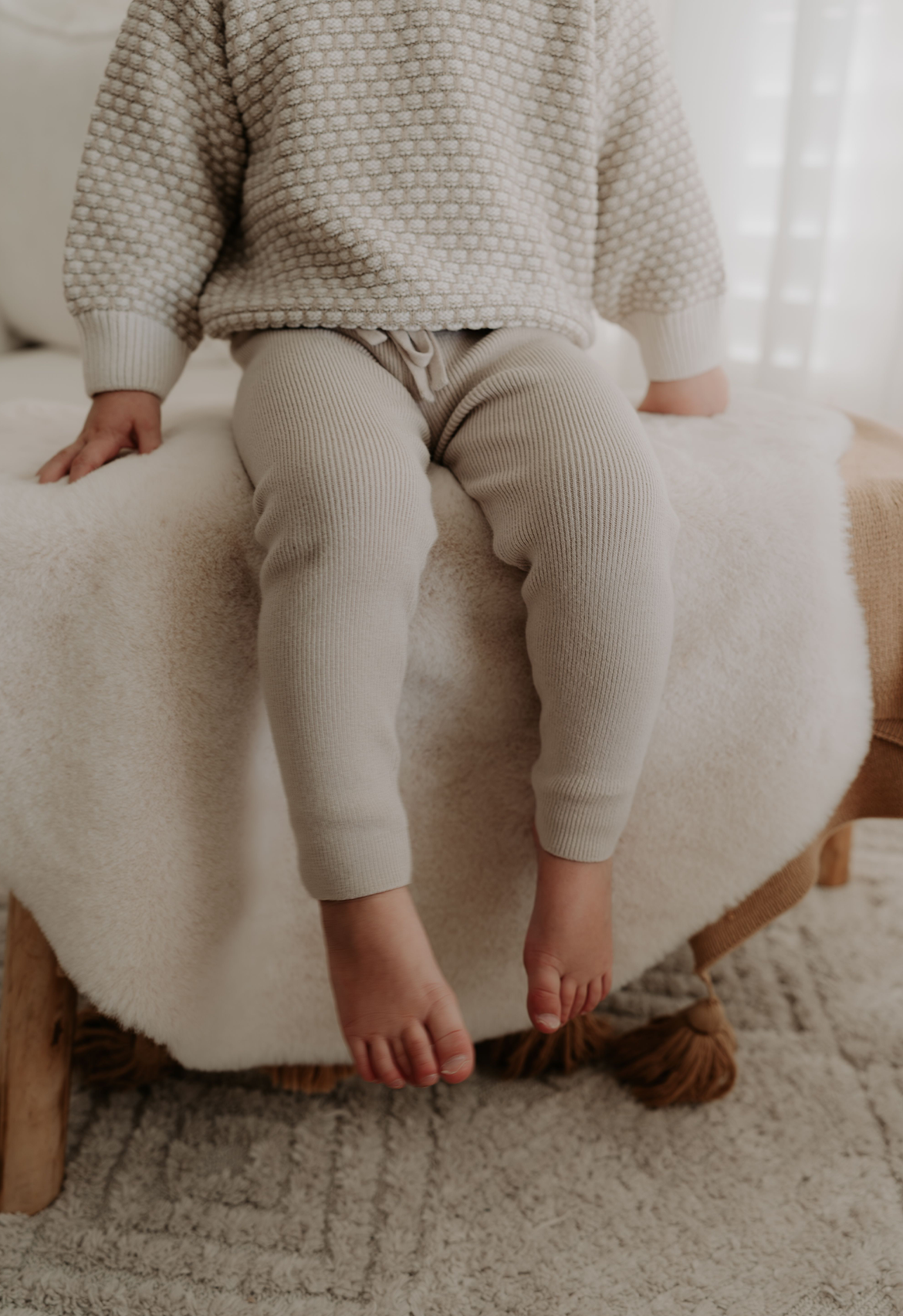Child wearing a textured sweater and light-coloured legging sitting on a white ottoman.