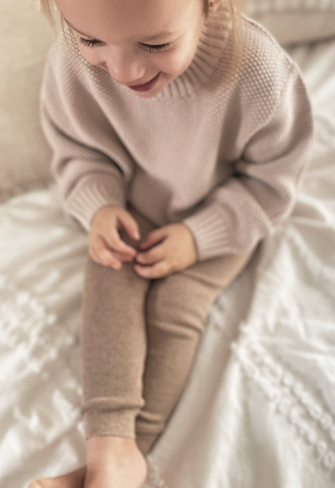A child sitting and smiling, wearing a beige chunky knit jumper and matching leggings.