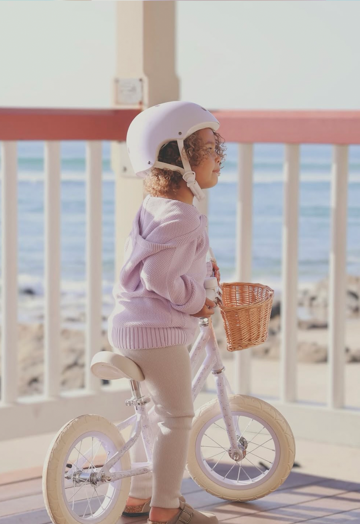 Child wearing a helmet and light purple sweater, standing next to a white bicycle with a basket on a balcony.
