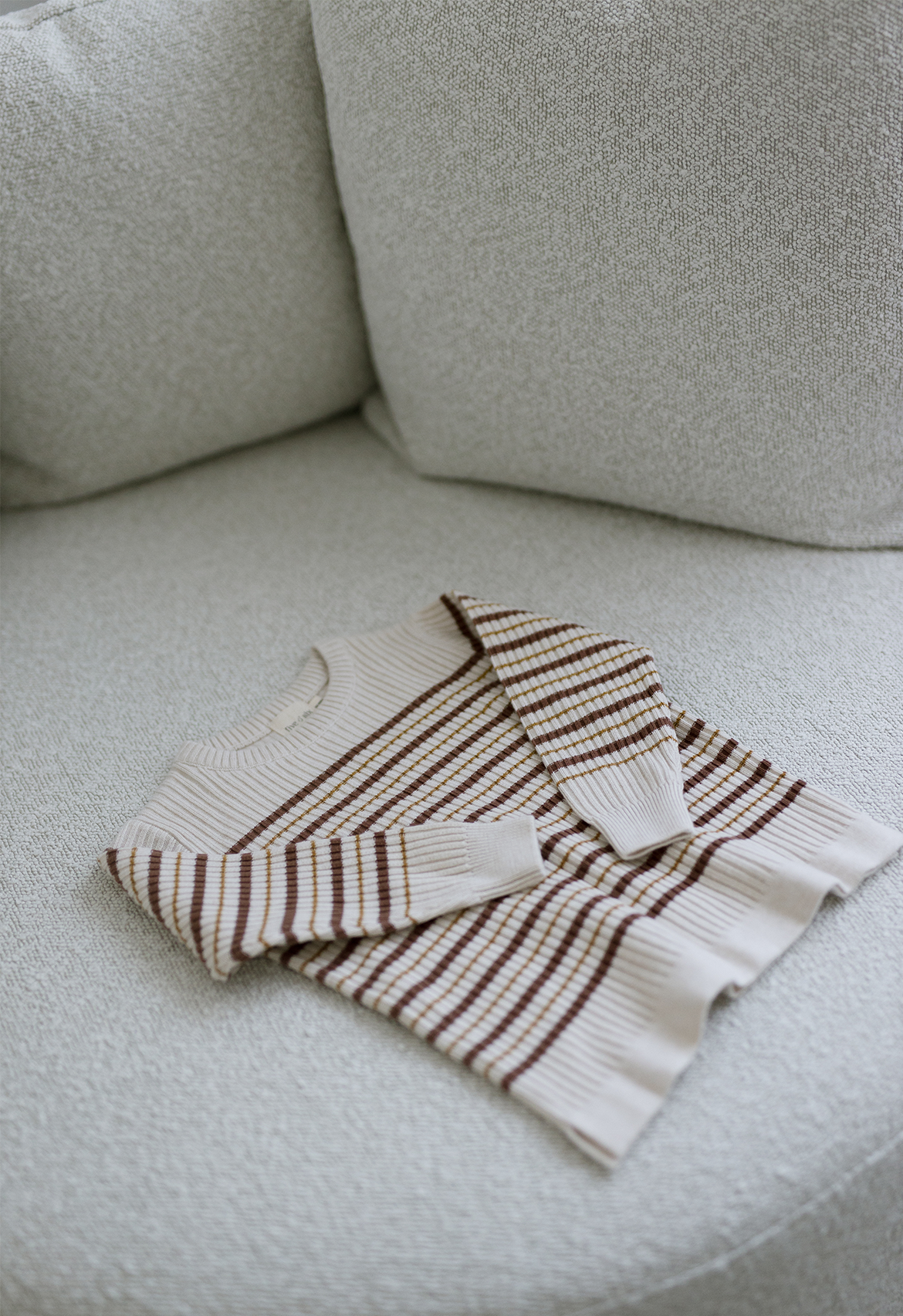 A striped ribbed jumper laid flat on a neutral colored surface, displaying an oatmeal base with russet and mustard stripes.
