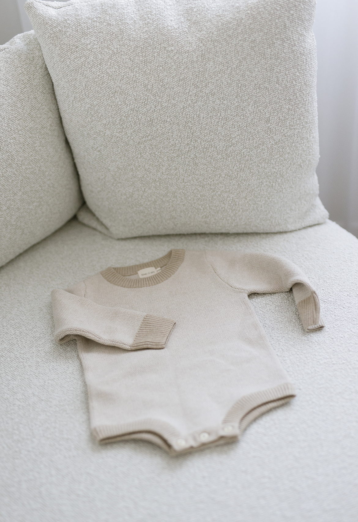 A long-sleeved beige baby romper with a round neckline, laid flat on a white couch