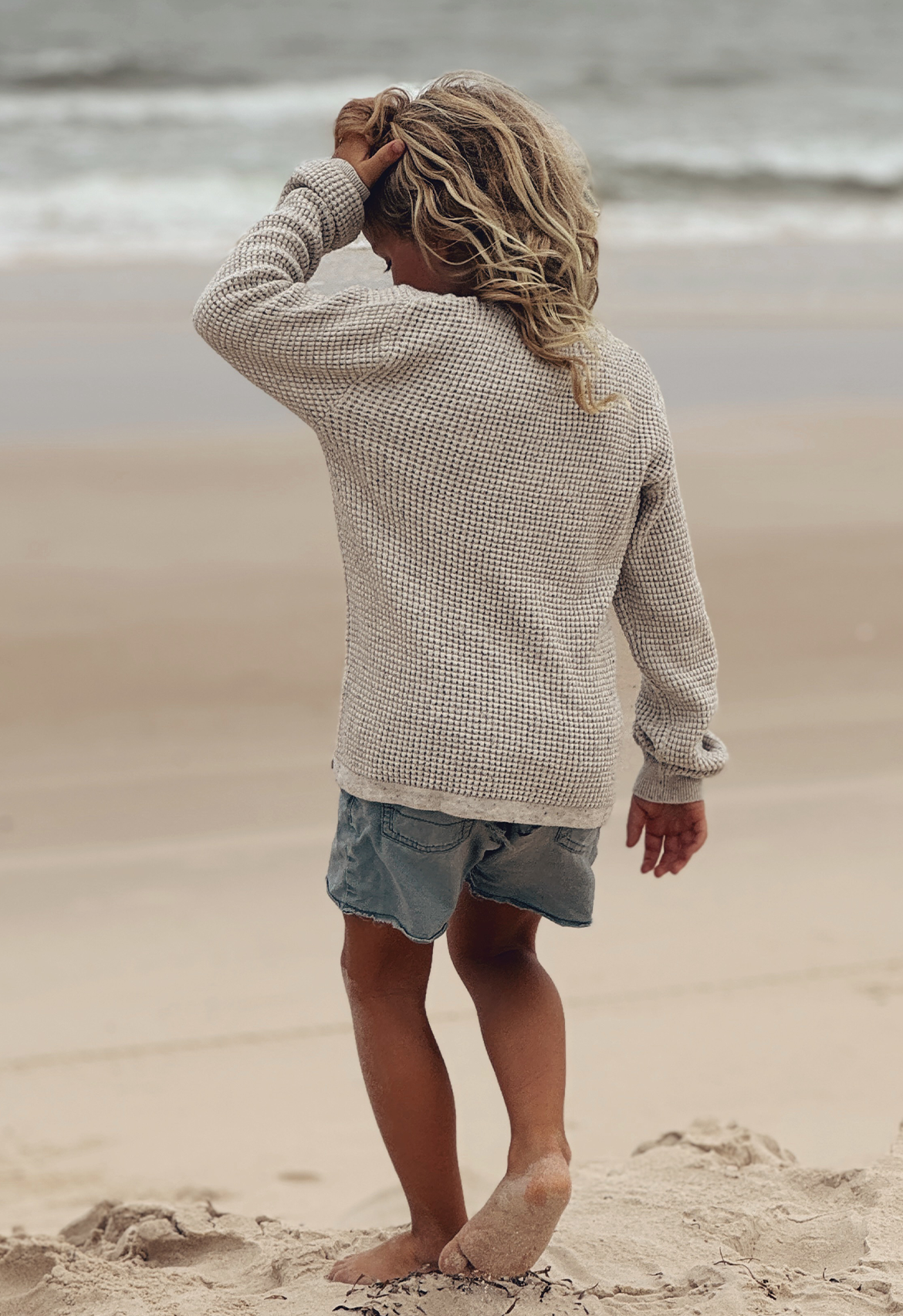 A child standing on a beach wearing an ivory speckle waffle texture jumper and denim shorts.